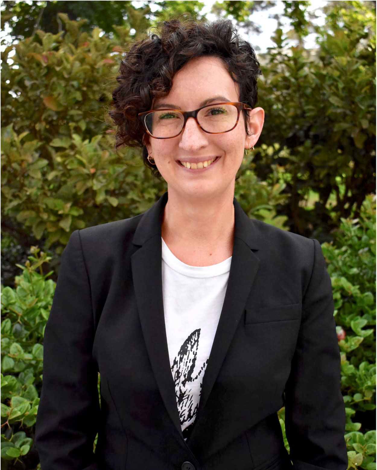 Cressida Madigan wearing a white shirt in a black blazer and glasses, standing in front of a tree.